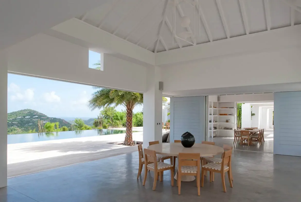 A round wooden dining table with woven chairs and a glossy black spherical vase sits beneath vaulted white beamed ceilings. Light blue doors open to an infinity pool that dissolves into lush tropical hills and a bright Caribbean sky—dining at its most spectacular.