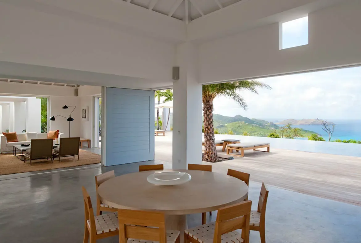 A round light wood dining table with woven chairs anchors the foreground while a white sofa and brown armchairs sit beyond. Floor-to-ceiling arches open to the infinity pool merging with the blue ocean and lush green hills—every meal comes with an unforgettable view.