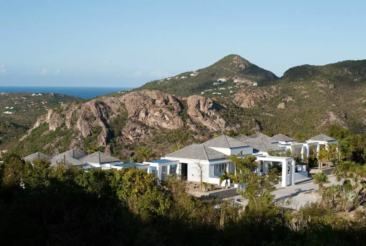 White contemporary pavilions with gray-shingled roofs nestle into lush tropical gardens at the foot of rugged brown hills. Beyond the mountains, the deep blue ocean stretches to the horizon under a brilliant cloudless sky—a private estate where architecture and nature exist in perfect harmony.