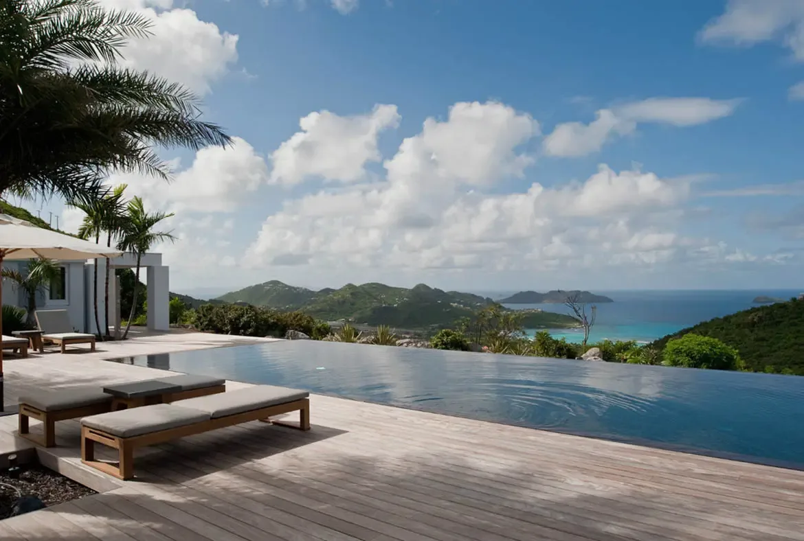 An infinity pool with crystal blue water merges seamlessly with rolling green hills and a turquoise ocean where distant islands float on the horizon. Wooden lounge chairs with plush cushions line the deck beneath palm fronds—where the pool ends and paradise begins is impossible to tell.