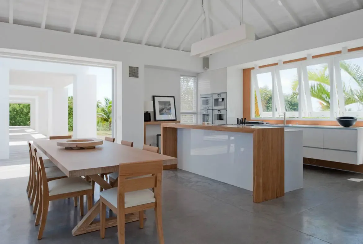 A sleek white kitchen island with a glossy countertop and warm wood panels sits beneath a long pendant light and vaulted white beams. Large windows frame palm trees and tropical greenery while a natural wood dining table anchors the foreground—cooking surrounded by nature.