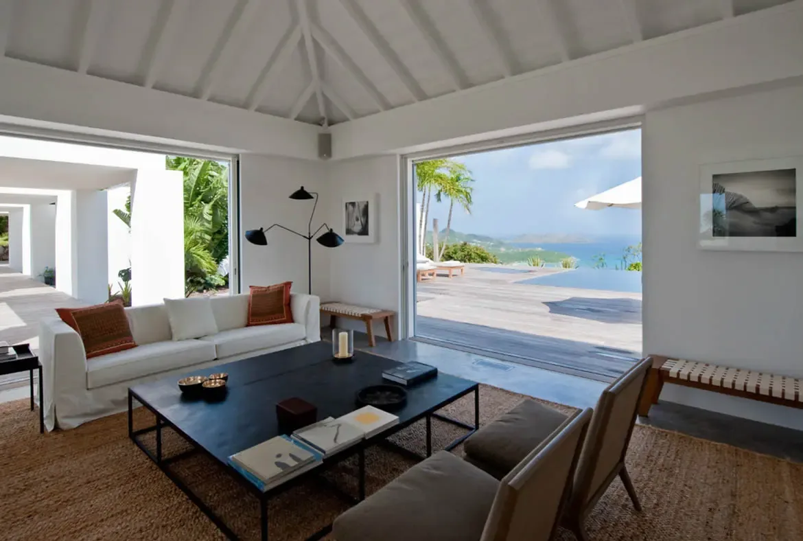 A white sofa with rust-colored patterned pillows and a dark coffee table with decorative bowls sit on a natural jute rug. The open arches frame an infinity pool and the blue ocean stretching to distant mountains—every angle of this living room is a postcard.