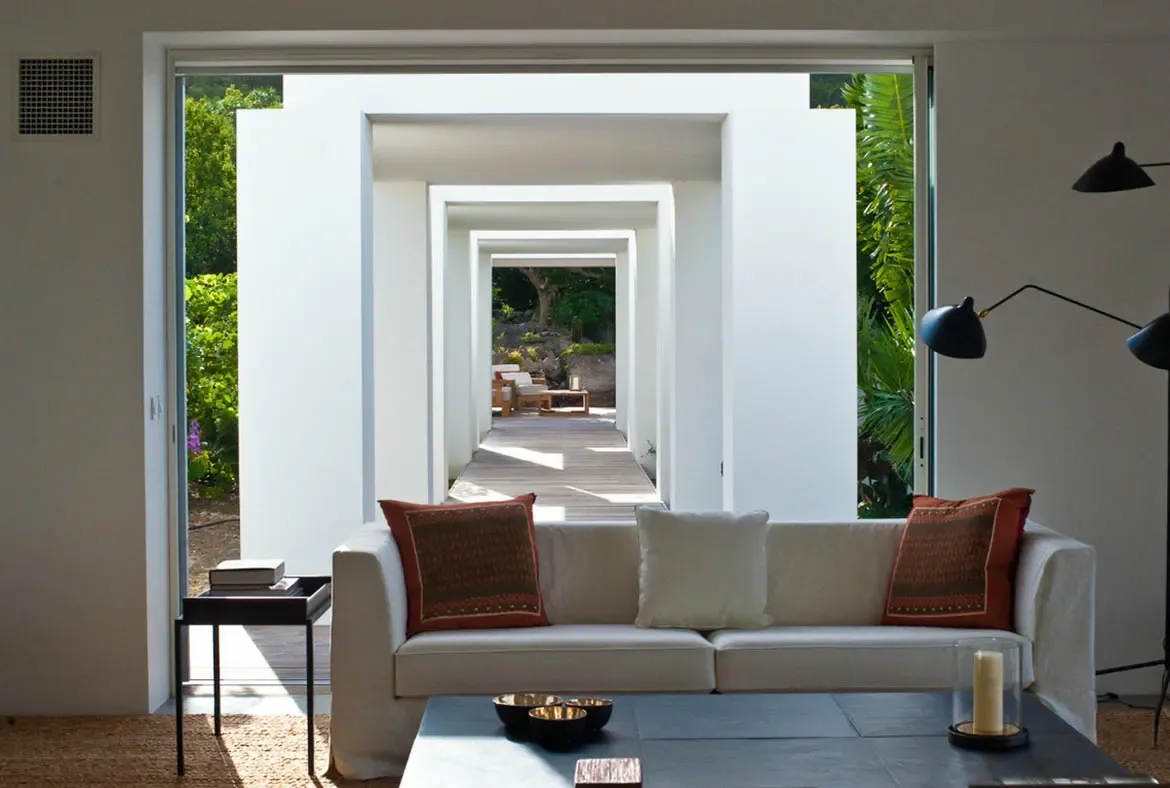 A cream sofa with earthy orange pillows sits on a natural jute rug before a series of nested white arches that tunnel toward lush tropical greenery. A black multi-arm floor lamp and dark coffee table with gold-interior bowls anchor the space—minimalist design that frames nature like art.