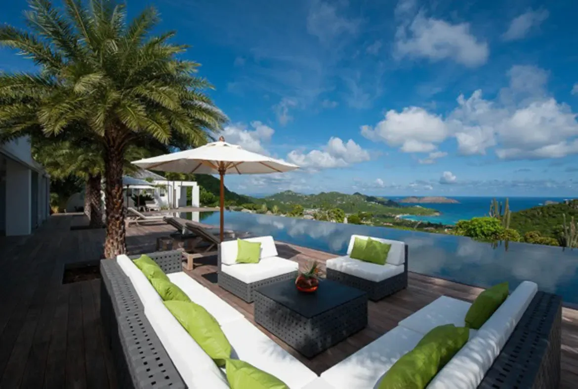 Dark wicker sofas with white cushions and lime-green throw pillows create a vibrant lounge on the wooden deck. An infinity pool dissolves into turquoise waters and distant islands while a palm tree sways overhead—outdoor living at its Caribbean finest.