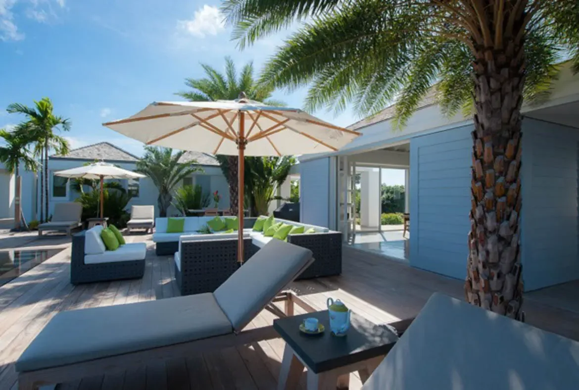 Light gray lounge chairs and a dark wicker sectional with vibrant green throw pillows sit beneath a large beige umbrella on a sun-drenched wooden deck. A blue teapot rests on the side table while tall palms sway overhead—afternoon relaxation perfected.