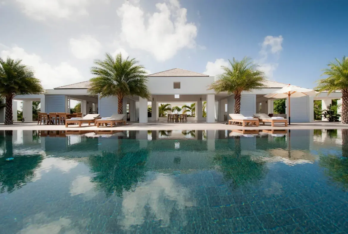 A turquoise pool mirrors the sky and palm trees beside a crisp white villa with open-air patios and light blue accents. Wooden lounge chairs and white umbrellas line the poolside while outdoor dining tables invite long alfresco lunches—your private Caribbean oasis awaits.