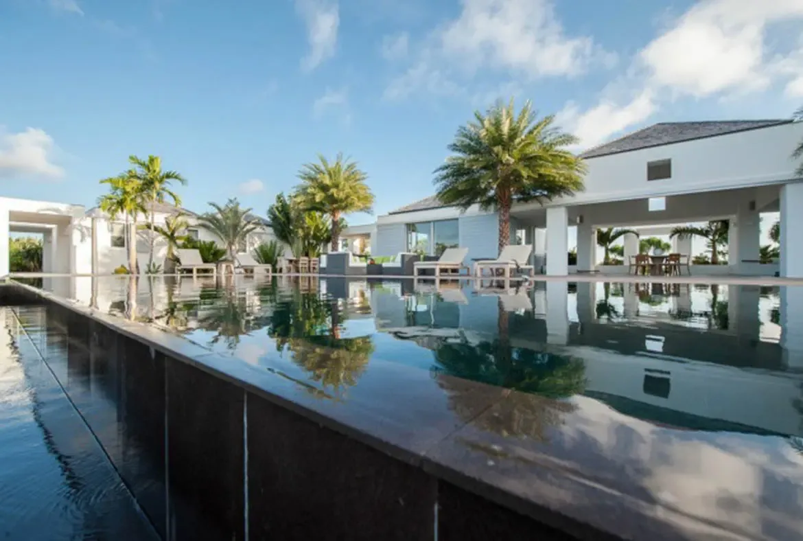 The crystal pool mirrors the blue sky and swaying palms beside sleek white contemporary architecture with open-air columns. White lounge chairs and a covered dining patio invite endless relaxation—this is the view that sells the dream.