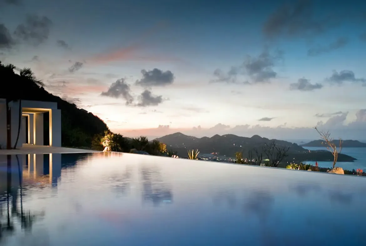 The infinity pool mirrors a sunset sky painted in soft orange and pink as twilight settles over lush green hills and distant ocean. Warm light spills from the villa's illuminated columns while a solitary palm silhouette adds drama—golden hour at its most magical.