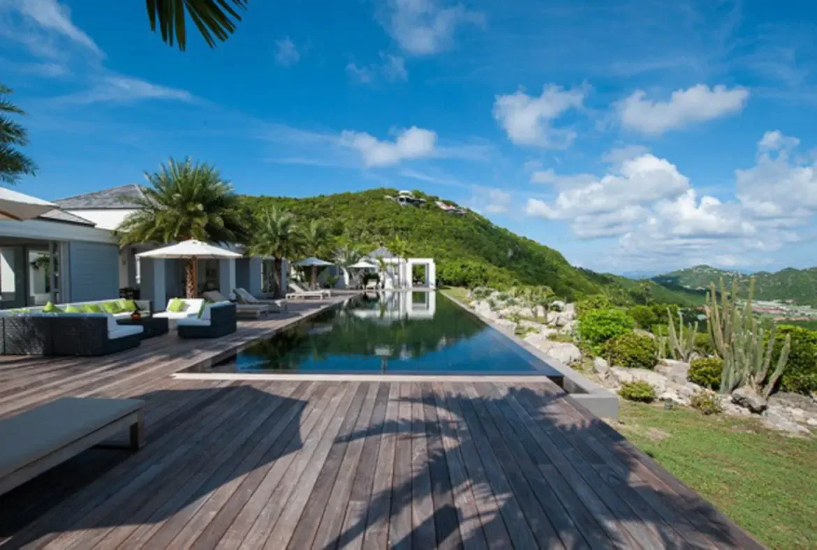 Dark wicker sofas with vibrant green cushions line the wooden deck beside a long sleek infinity pool that mirrors the brilliant blue sky. White umbrellas shade the loungers while palm shadows dance across the planks and rolling mountains fade into the horizon—endless Caribbean luxury.