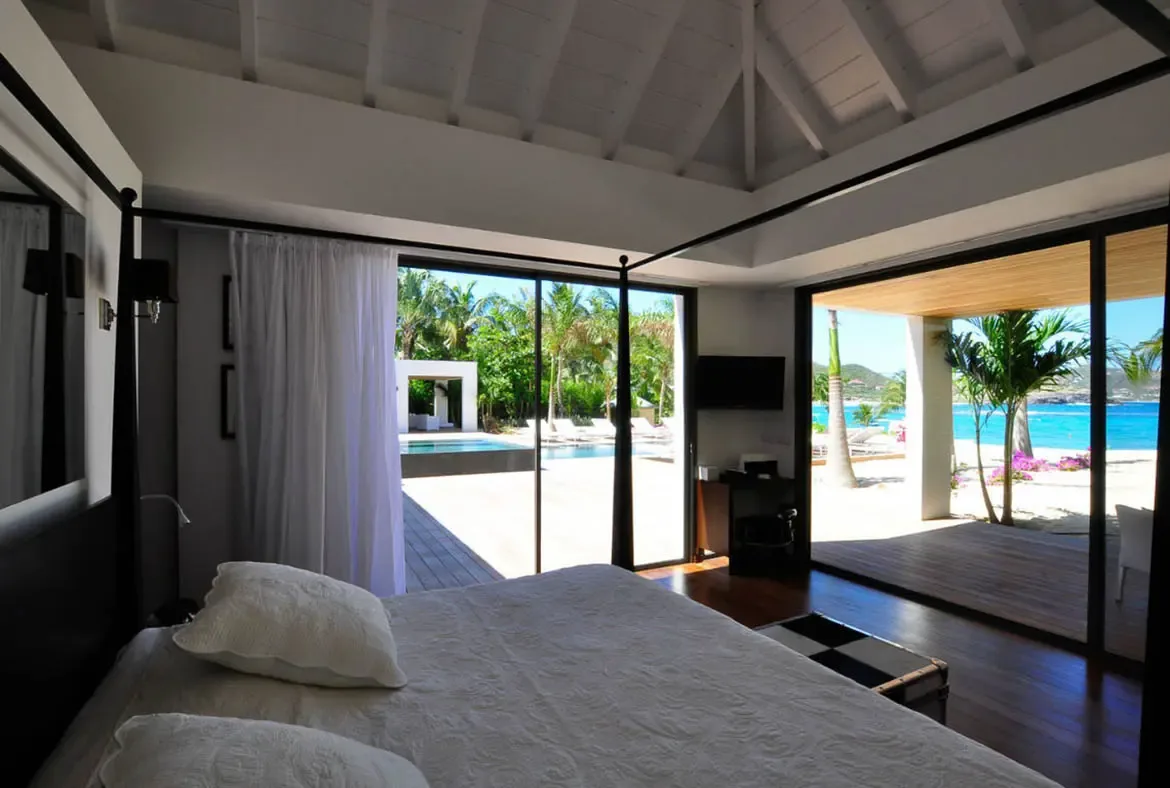 A white dining table with modern chairs sits before open glass walls, where a deep blue ocean panorama stretches to the horizon beneath wispy clouds.