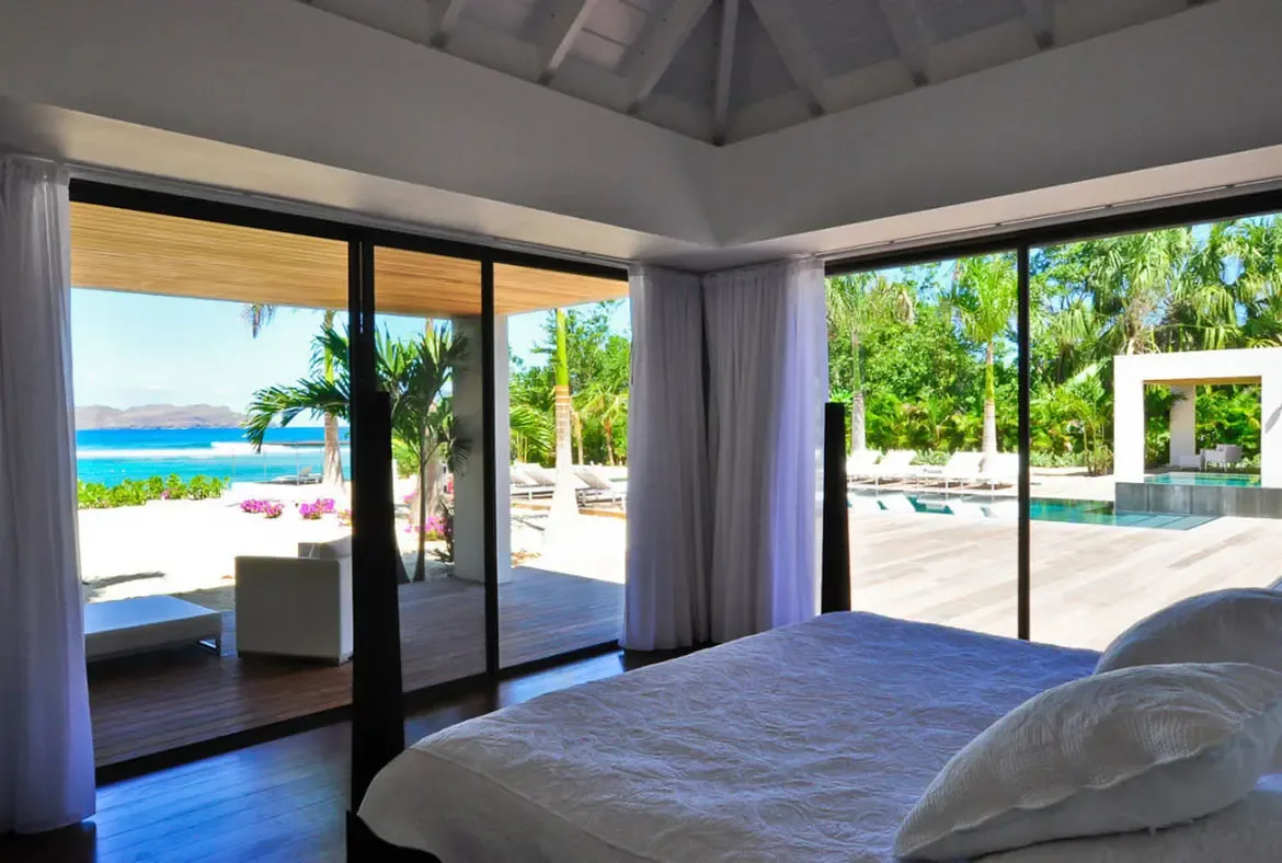 A white sectional sofa sits before floor-to-ceiling glass walls, where an infinity pool and turquoise ocean fill the minimalist living room with natural light.