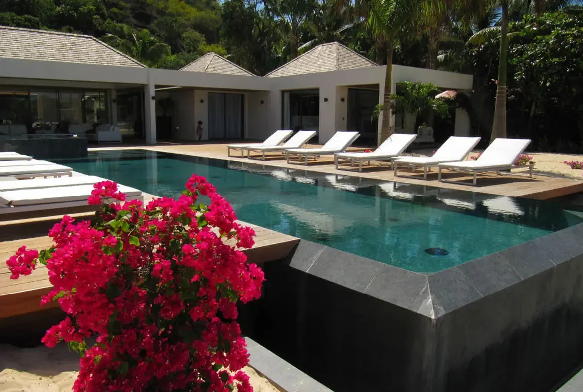White loungers beside an infinity pool sit on a spacious deck, where a white cabana and palm trees frame a turquoise ocean beneath puffy cumulus clouds.