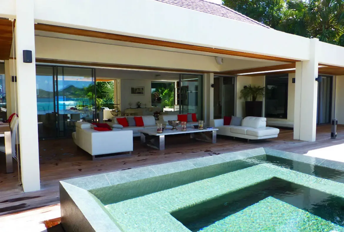 A blue-lit infinity pool reflects a white villa at night, where palm trees and warm interior lights create an elegant evening atmosphere against a dark hillside.