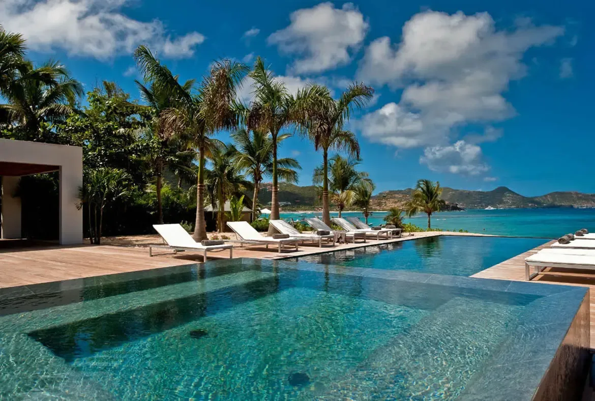 An infinity pool reflects golden light at sunset, where white loungers and a modern white villa glow against a pastel sky and silhouetted hills.