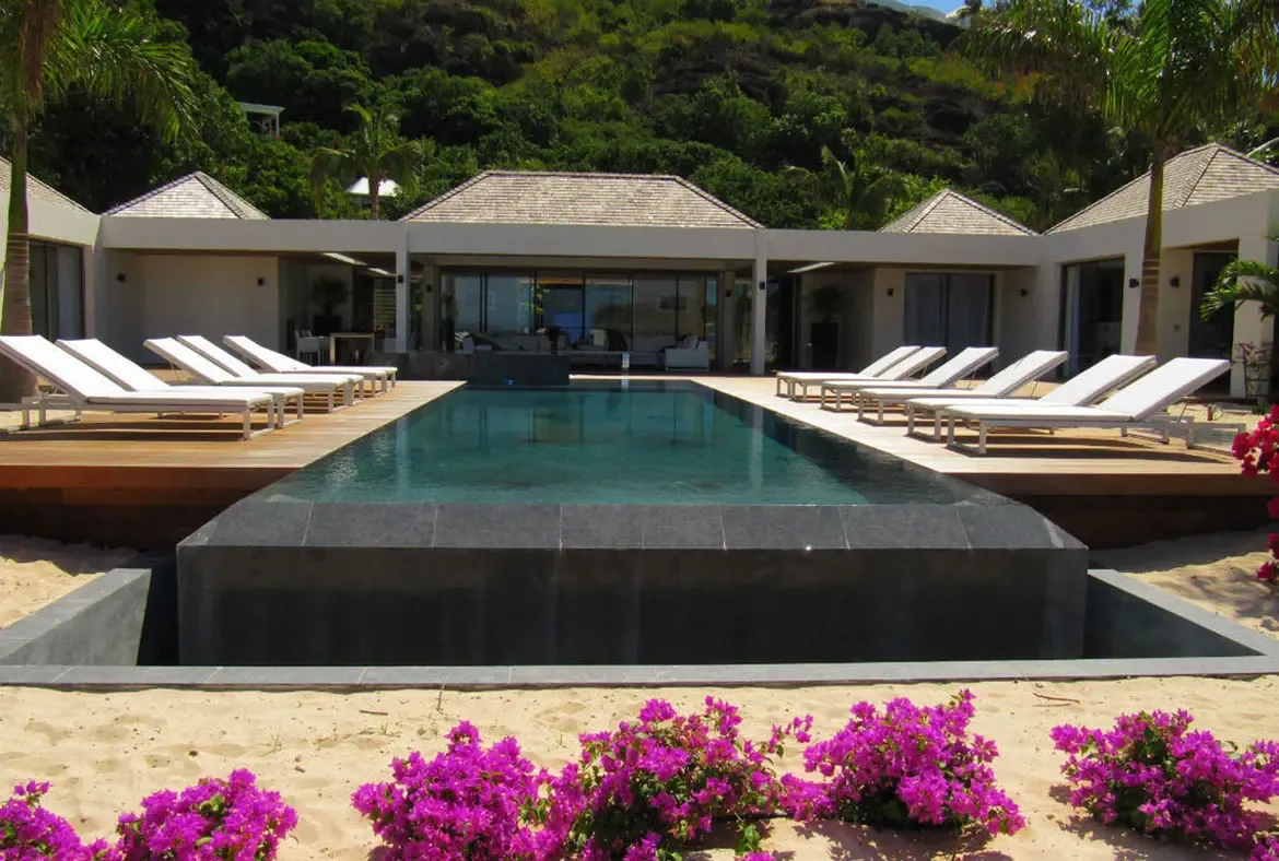An outdoor sofa with colorful pillows sits on a covered terrace, where white pillars and lush tropical greenery frame a peaceful garden retreat.