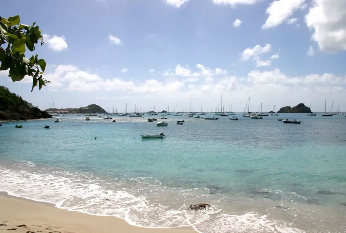 Pale golden sand meets crystal-clear turquoise water where gentle waves create white foam along the shore. Sailboats and yachts anchor peacefully in the bay beneath rolling green hills—a pristine Caribbean beach steps from the villa.
