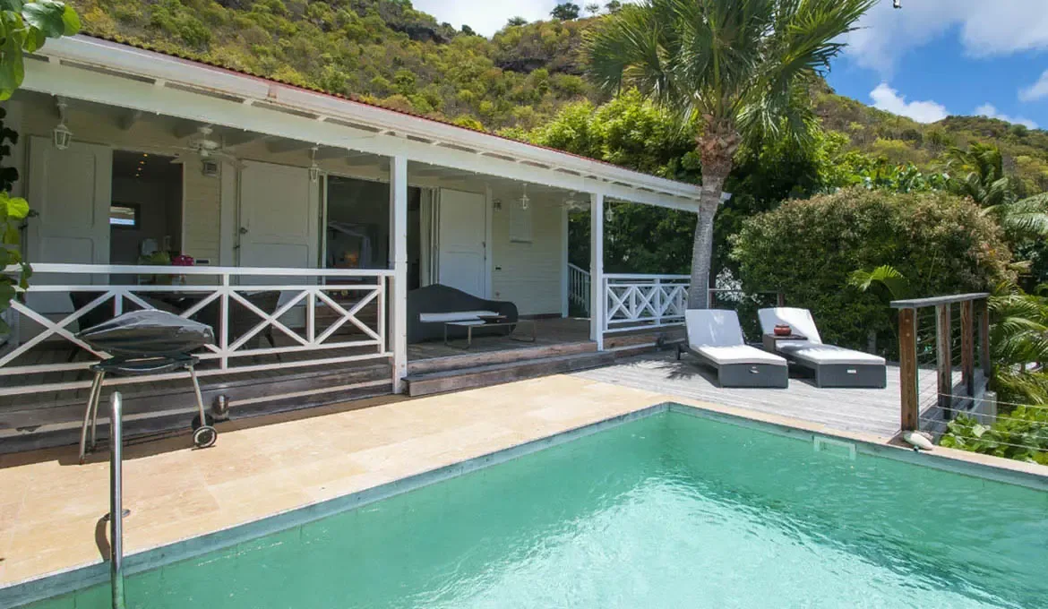 A white villa with a red-tiled roof and French doors opens onto a covered porch with white X-design railings. A turquoise pool glistens on the stone terrace beside white lounge chairs and a barbecue grill while lush green hills rise behind—your private hillside retreat in St. Barths.