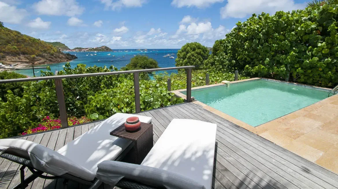 Two white lounge chairs with gray cushions sit on a weathered wooden deck beside a turquoise pool framed by pink blooms. Beyond the railing, sailboats and yachts dot a calm bay beneath rolling green hills—your private terrace above the Caribbean fleet.