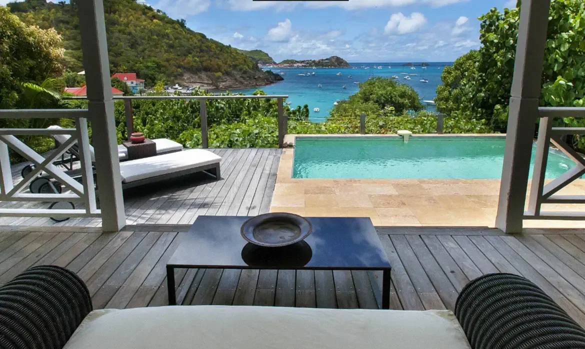 A dark wooden deck with a sleek black coffee table sits beside a turquoise pool on a stone terrace framed by white X-design railings. Below the hillside, yachts dot a vibrant blue bay while red-roofed buildings peek through lush green vegetation—terrace living at its finest.