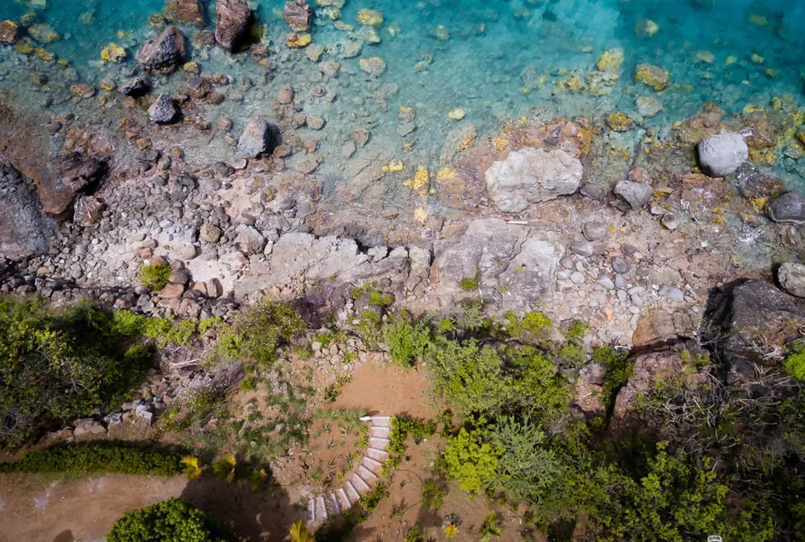 Crystal turquoise water reveals submerged rocks and coral patterns along a rugged shoreline where massive boulders meet the Caribbean sea. A winding path through lush greenery leads to light-colored steps descending to the water—a bird's-eye view of paradise at your doorstep.