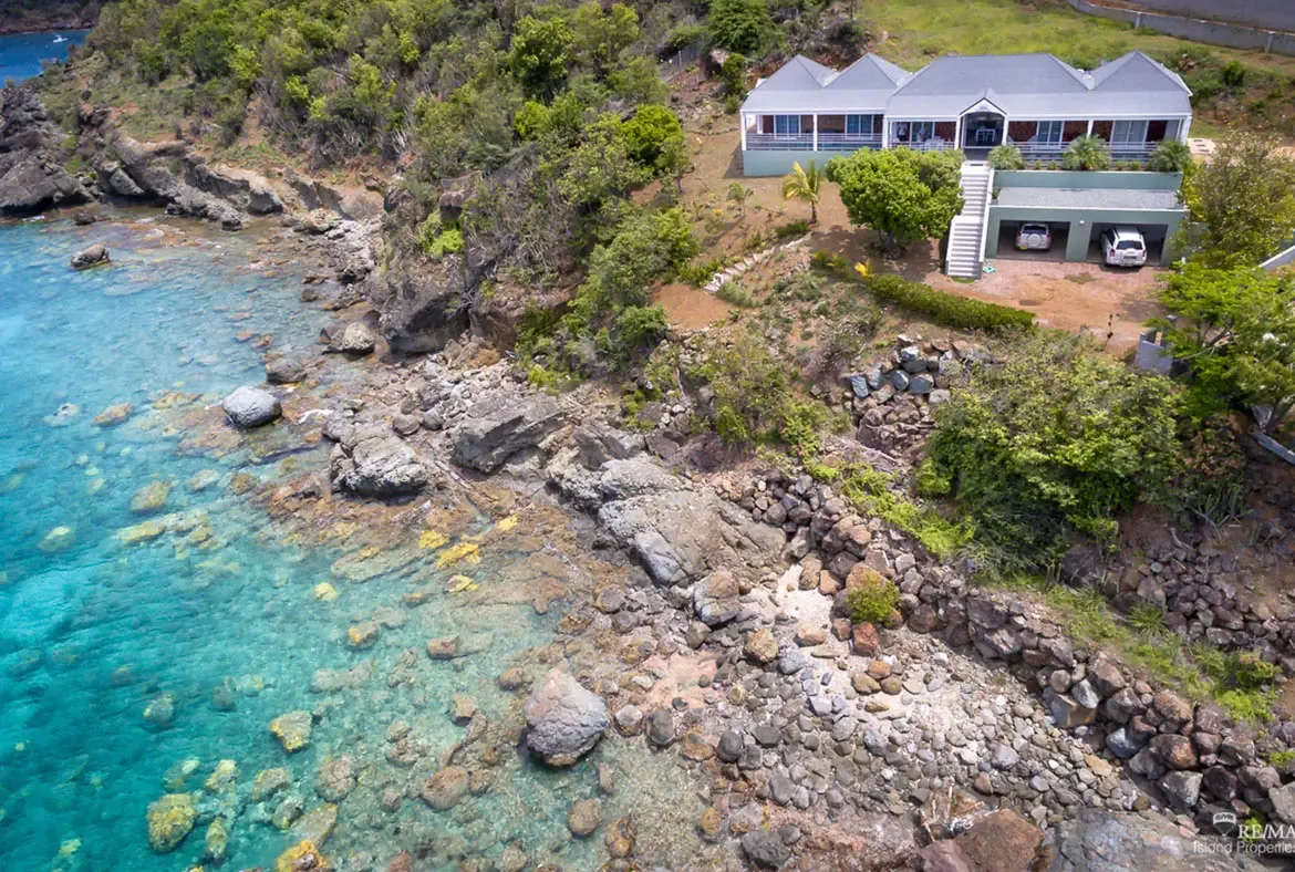 An aerial view reveals a modern villa with a gray gabled roof perched on a lush hillside above a rocky turquoise coastline. A white staircase descends through tropical greenery while two vehicles park beside the garage—the ultimate private Caribbean retreat from above.