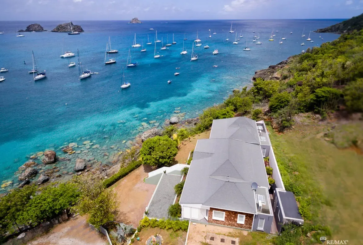 An aerial panorama shows the modern villa with its gray gabled roof and white siding perched above a bay where dozens of sailboats and yachts anchor in crystal turquoise water. Rocky outcrops and lush tropical greenery frame the coastline while distant islets dot the horizon—your private hillside overlooking the Caribbean fleet.