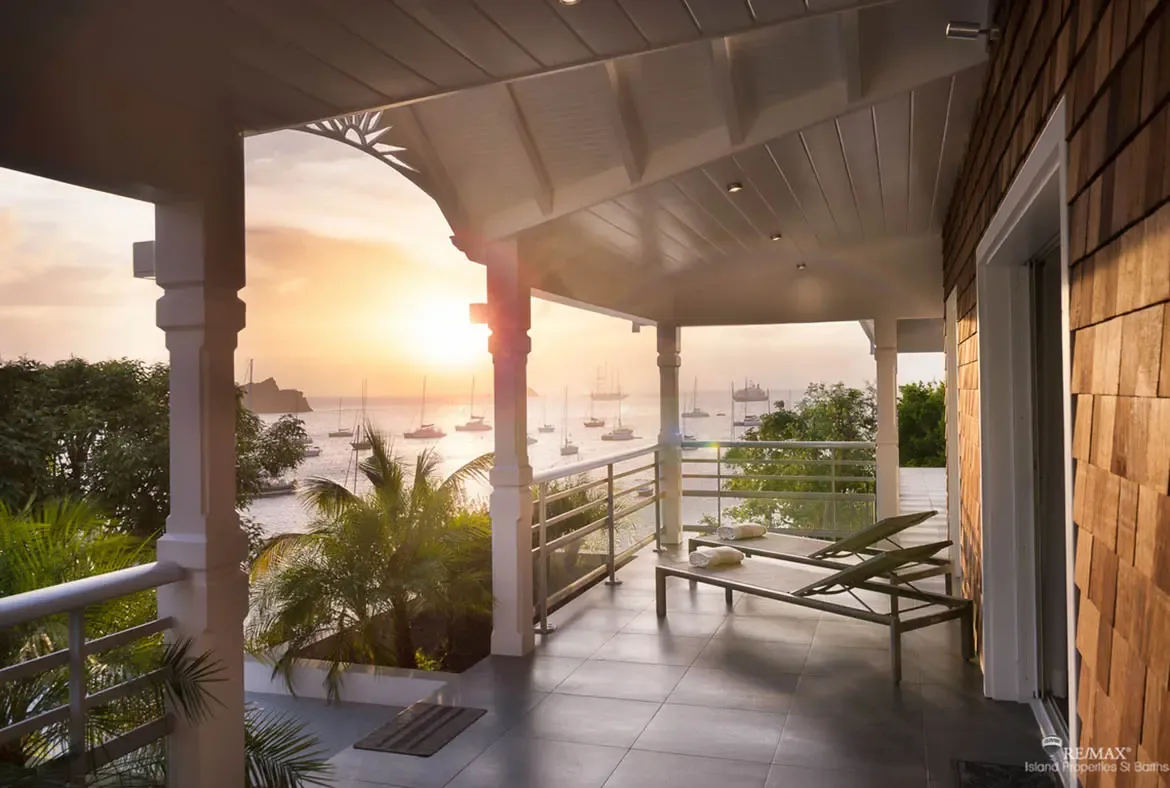 Two lounge chairs with rolled white towels sit beneath ornate white columns as golden sunset light bathes the balcony. Yachts and sailboats dot the tranquil ocean beyond lush tropical palms while wooden shingles add warmth—sunset cocktails are calling.