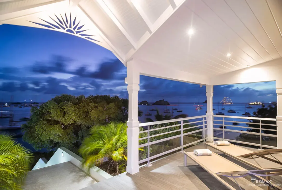 Two beige lounge chairs with white towels sit beneath a white vaulted ceiling with a sunburst cutout as twilight settles over a calm harbor. Illuminated yachts anchor beyond the tropical shoreline while the deepening sky casts a moody blue glow—evenings on this balcony are unforgettable.