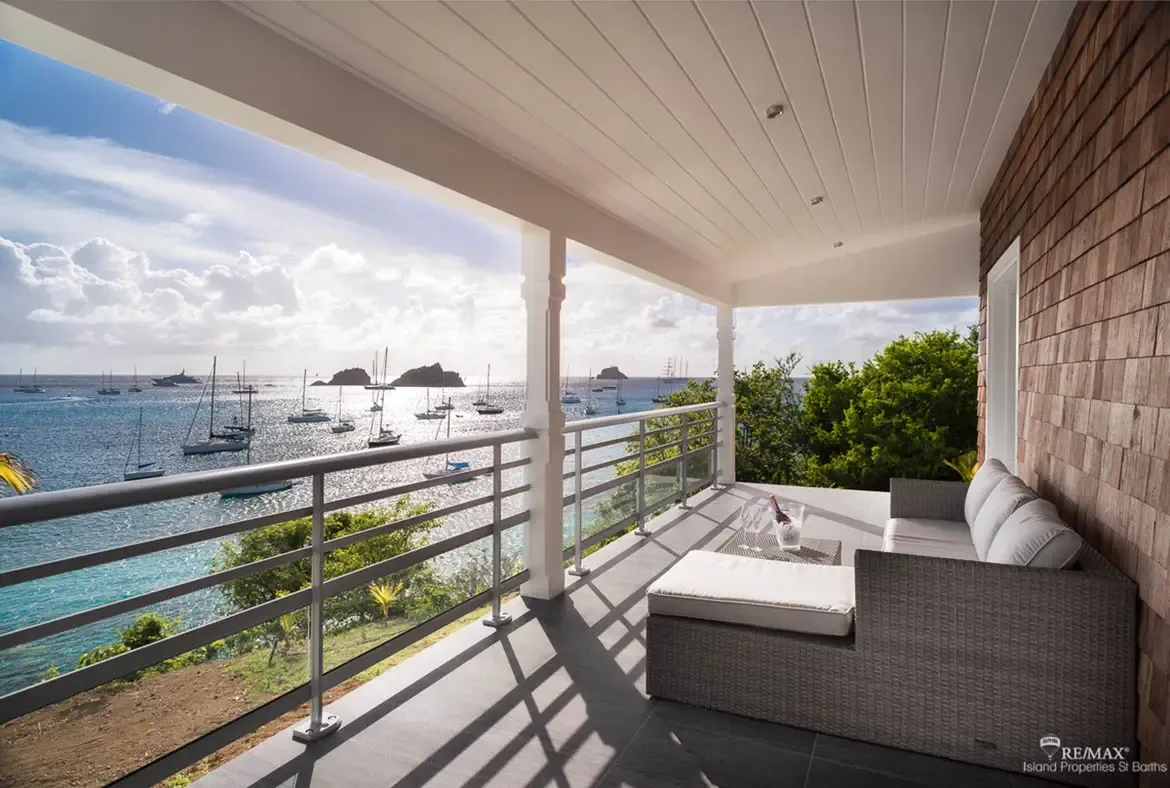 Woven wicker chairs with plush white cushions sit beside a small table holding wine glasses and a bottle. Beyond the metal railing, sailboats and yachts anchor in crystal turquoise water near rocky islets while the late afternoon sun casts warm golden light—happy hour perfection.