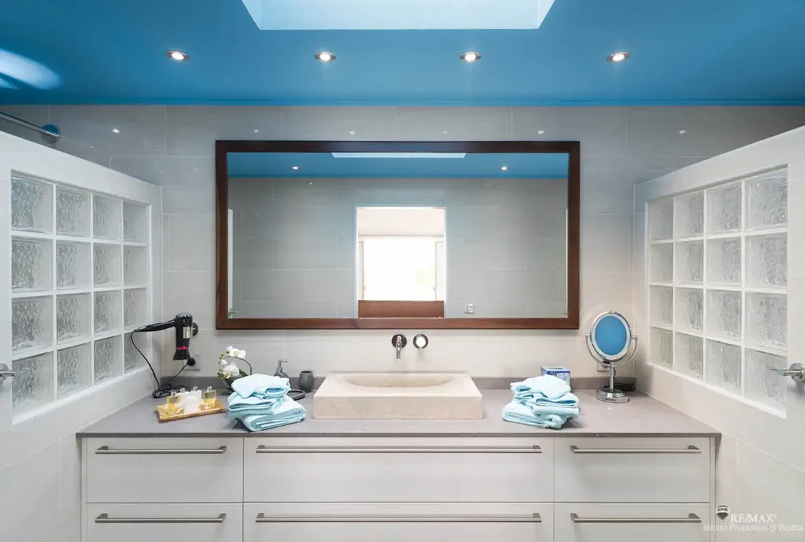 A bold bright blue ceiling with a skylight floods this bathroom with natural light. Frosted glass block panels frame the shower while a vessel sink and six-drawer vanity sit below a dark-framed mirror—white orchids and light blue towels add the finishing touches to a bathroom that feels like the Caribbean sky.