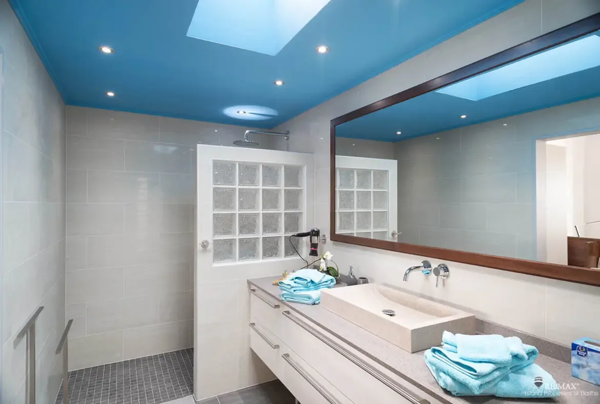 A vibrant blue ceiling with a skylight crowns this double-vanity bathroom featuring two vessel sinks on a light stone countertop. Frosted glass block walls enclose the rain shower while light blue towels echo the ceiling—twin sinks, twin views of paradise.