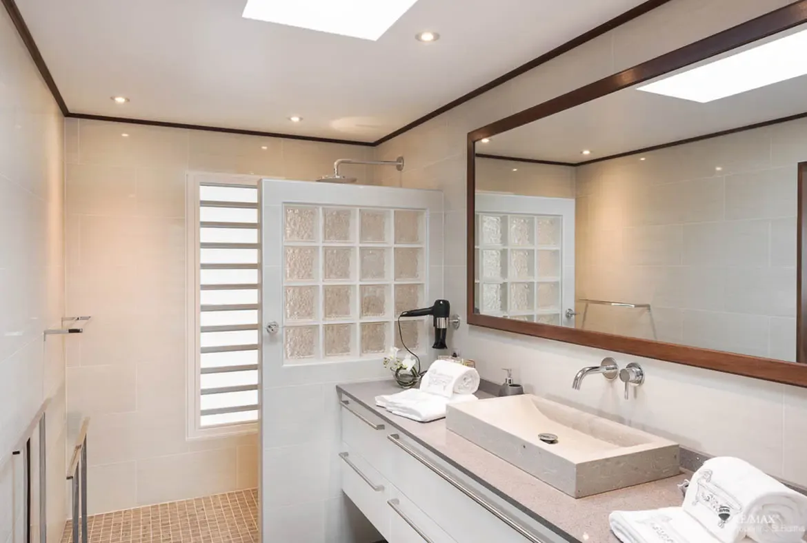 A frosted glass block shower wall diffuses natural light beside a window with horizontal slats. A rectangular vessel sink with white flowers sits below a dark-framed floor-to-ceiling mirror while a skylight bathes the beige tile walls in sunshine—practical elegance meets Caribbean brightness.