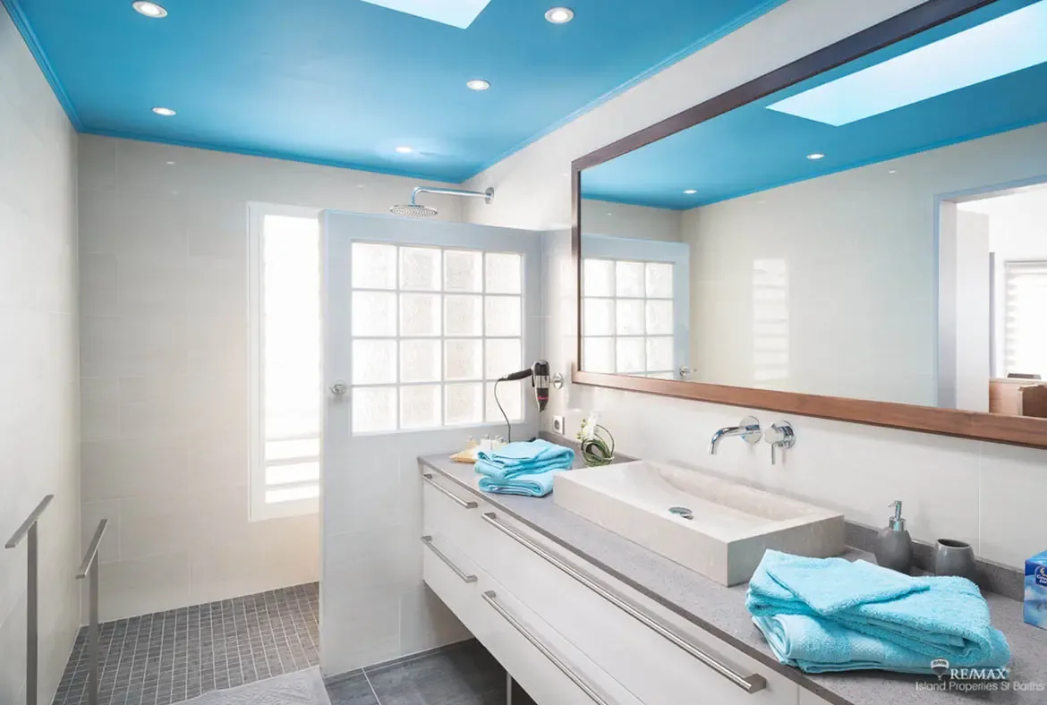 A turquoise-blue ceiling with recessed lighting crowns this modern bathroom with light gray tiled walls and a glass-block shower window. A vessel sink on a stone countertop sits below a dark-framed mirror while turquoise towels echo the bold ceiling color—a bathroom that makes a statement.