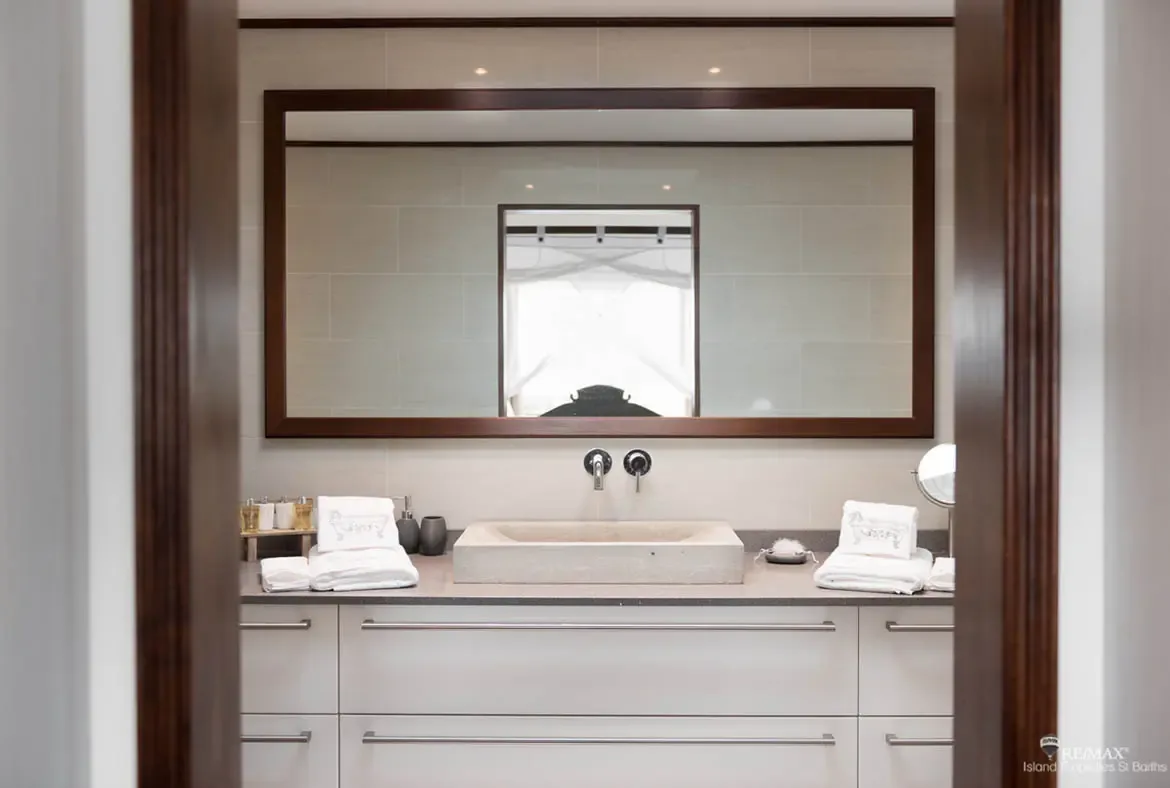A dark-framed rectangular mirror sits above a light stone vessel sink with sleek wall-mounted faucets. Neatly folded white towels and gray ceramic containers line the countertop while warm beige tiles and recessed lighting add sophistication—a bathroom with boutique hotel elegance.