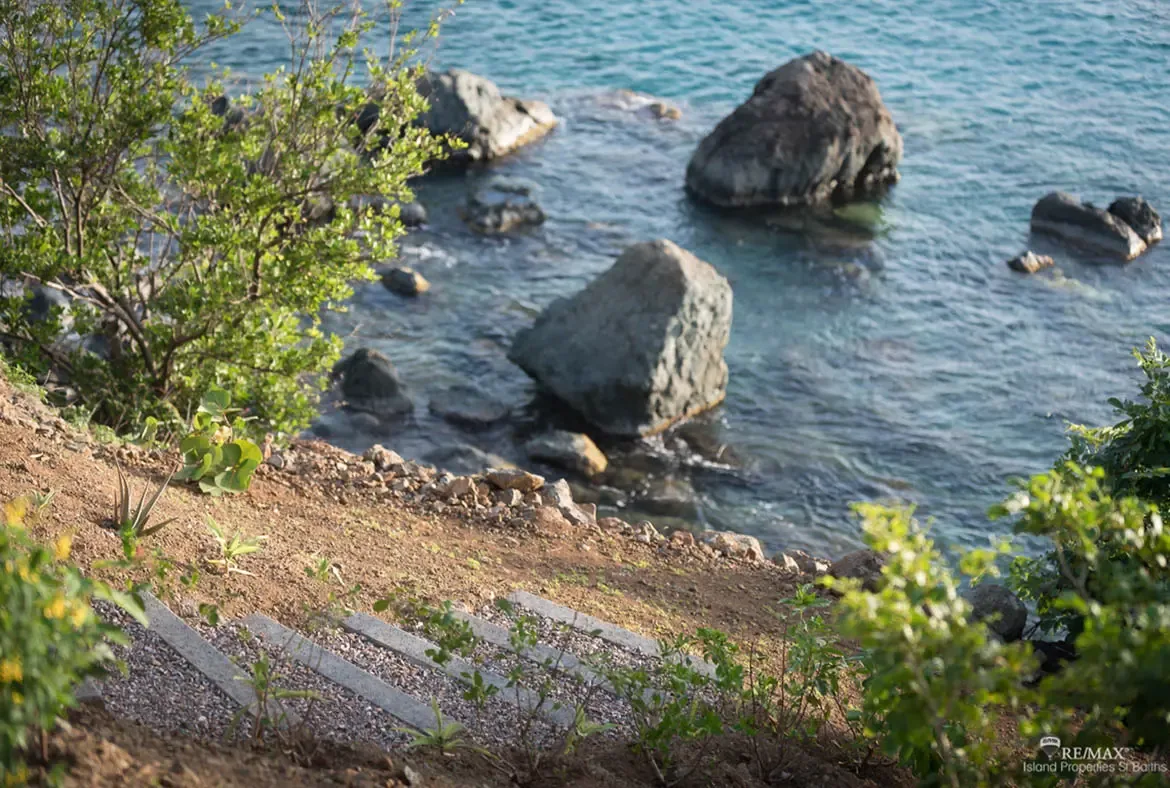 A stepping-stone path lined with wildflowers and lush greenery winds down to a rocky shoreline where weathered boulders meet crystal-clear turquoise water. Gentle ripples reflect sunlight on the calm Caribbean sea—your private path to the beach.