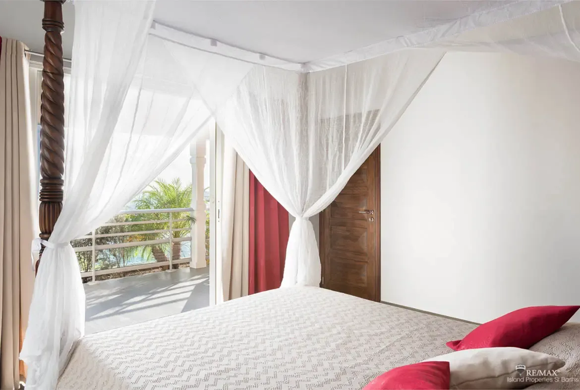 A four-poster bed draped with a sheer white canopy tied back with ribbons features red and white throw pillows on a light patterned spread. Bold red curtains frame the open doors to a sun-drenched terrace with swaying palms—sleep under canopy elegance in the heart of St. Barths.