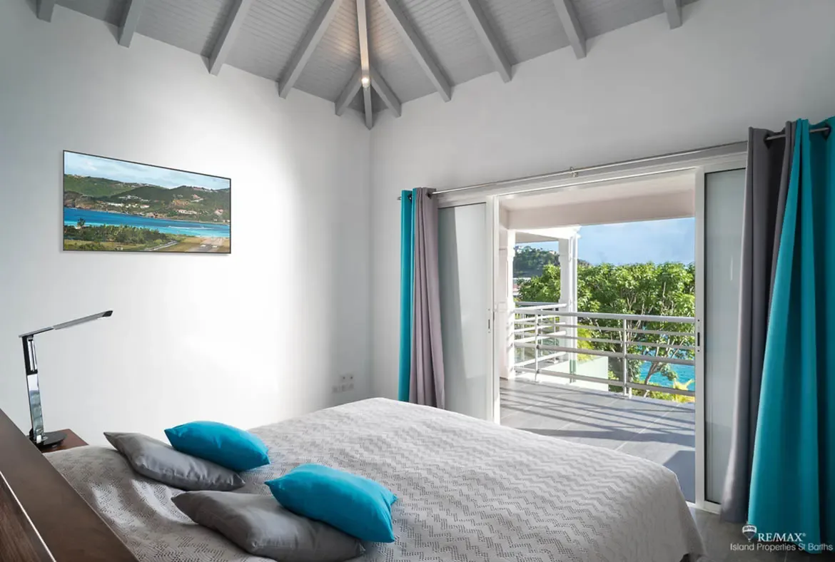 A light gray bedspread with vibrant turquoise pillows faces a large coastal landscape painting in matching hues. Turquoise and gray curtains frame open doors to a balcony overlooking lush greenery and the ocean beyond—the art and the view are one and the same.