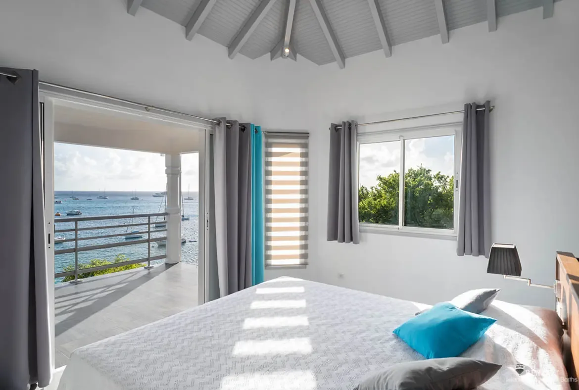 A white-patterned bedspread with a vibrant turquoise accent pillow faces open doors leading to a balcony overlooking yachts on calm Caribbean water. Gray and turquoise curtains frame the vaulted beamed ceiling while lush greenery peeks through a side window—a bedroom that wakes up to paradise.