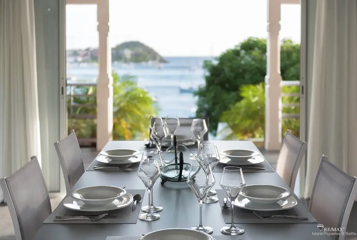 A dark dining table set for six with polished silverware and wine glasses sits beneath pale pink columns and sheer white curtains. Beyond the open archways, yachts anchor in a calm bay while a lush green island rises from the turquoise water—dining with a Caribbean masterpiece for a backdrop.