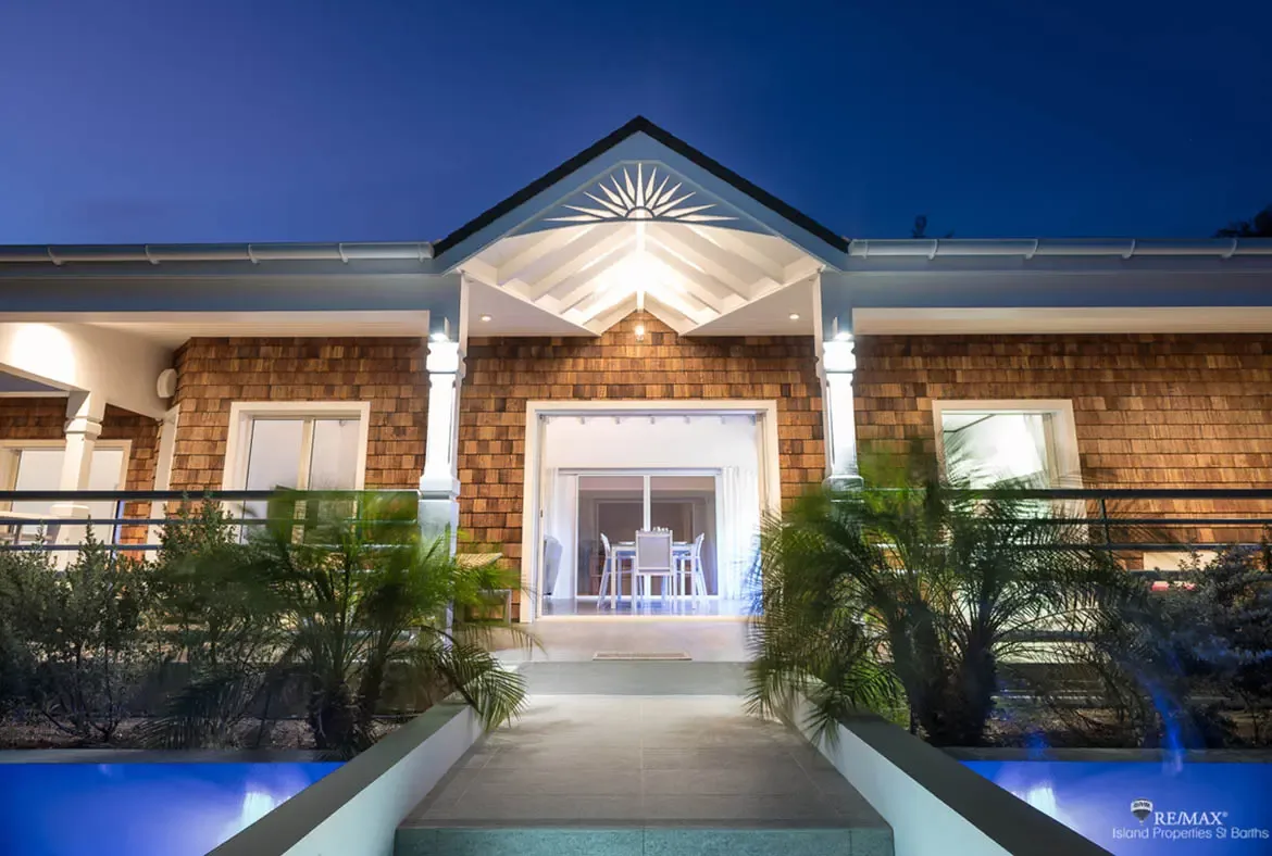 A gabled portico with a sunburst motif glows warmly above open doors revealing a bright dining interior. Rectangular blue-lit water pools flank the stone pathway while twin palm trees stand guard—arriving at Villa Pleasure feels like stepping into a dream.