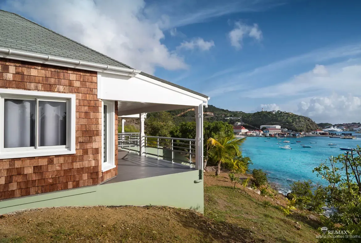 A warm-shingled villa with white columns and a covered porch perches on a hillside above a turquoise bay dotted with yachts. Across the water, red-roofed buildings cluster beneath lush green hills while a distant lighthouse crowns the view—hillside living with the whole Caribbean at your feet.