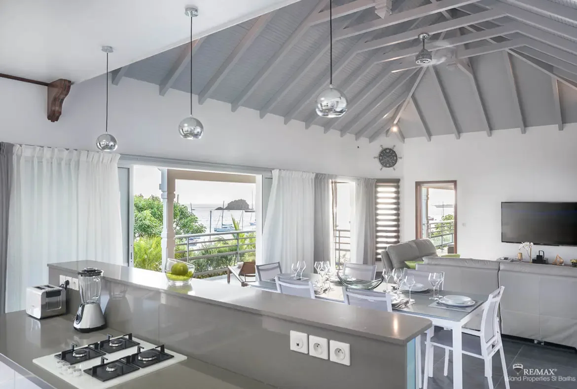 A gray kitchen island with a gas cooktop sits beneath three silver globe pendants and vaulted light-gray beams. A glass-topped dining table set with white plates and wine glasses leads to a cozy sectional facing the TV—open-concept living where the Caribbean ocean peeks through the curtains.