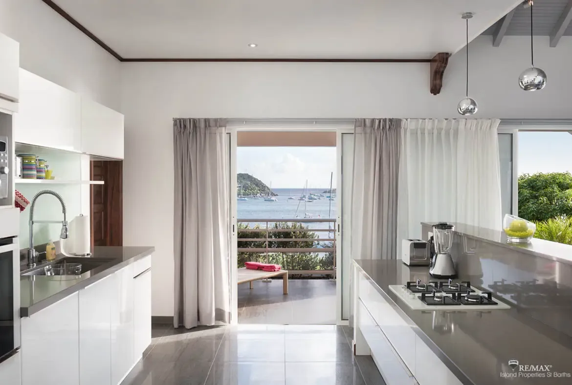 Sleek glossy white cabinetry and dark countertops anchor a modern kitchen where two sphere pendants hang above. Flowing curtains frame open doors to a balcony overlooking sailboats on turquoise water and a hilly island beyond—cooking with a Caribbean panorama.