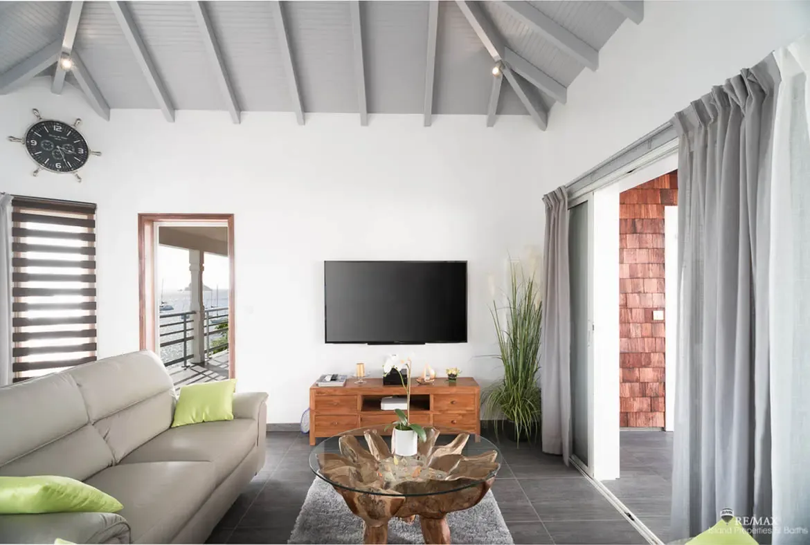 A light gray sofa with vibrant green throw pillows sits beside a glass coffee table with a driftwood-style base on a soft gray rug. Through the open doorway, boats drift on calm water while a nautical clock and white orchid add coastal charm—relaxed living meets Caribbean views.