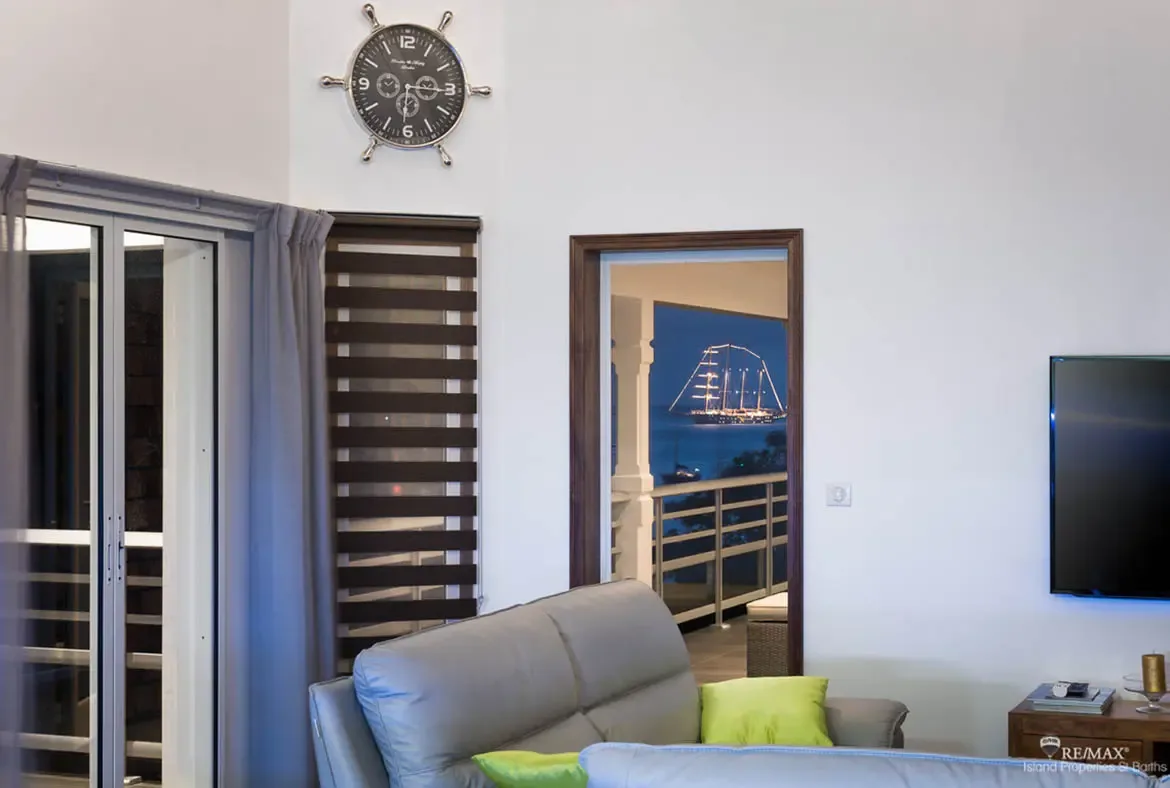 A light gray leather sofa with vibrant green cushions faces a flat-screen TV while a nautical ship's wheel clock anchors the coastal theme. Through dark-framed doors, a brightly lit sailboat glows on the nighttime ocean—evenings here are pure magic.