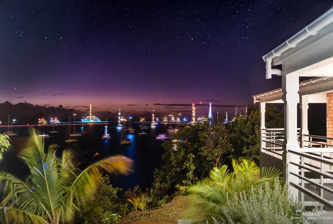 A starry sky transitions from deep indigo to soft purple above a calm harbor where dozens of illuminated yachts cast shimmering reflections on the water. Palm fronds frame the view from the villa balcony while warm interior lighting glows beside the railing—nights at Villa Pleasure are pure magic.