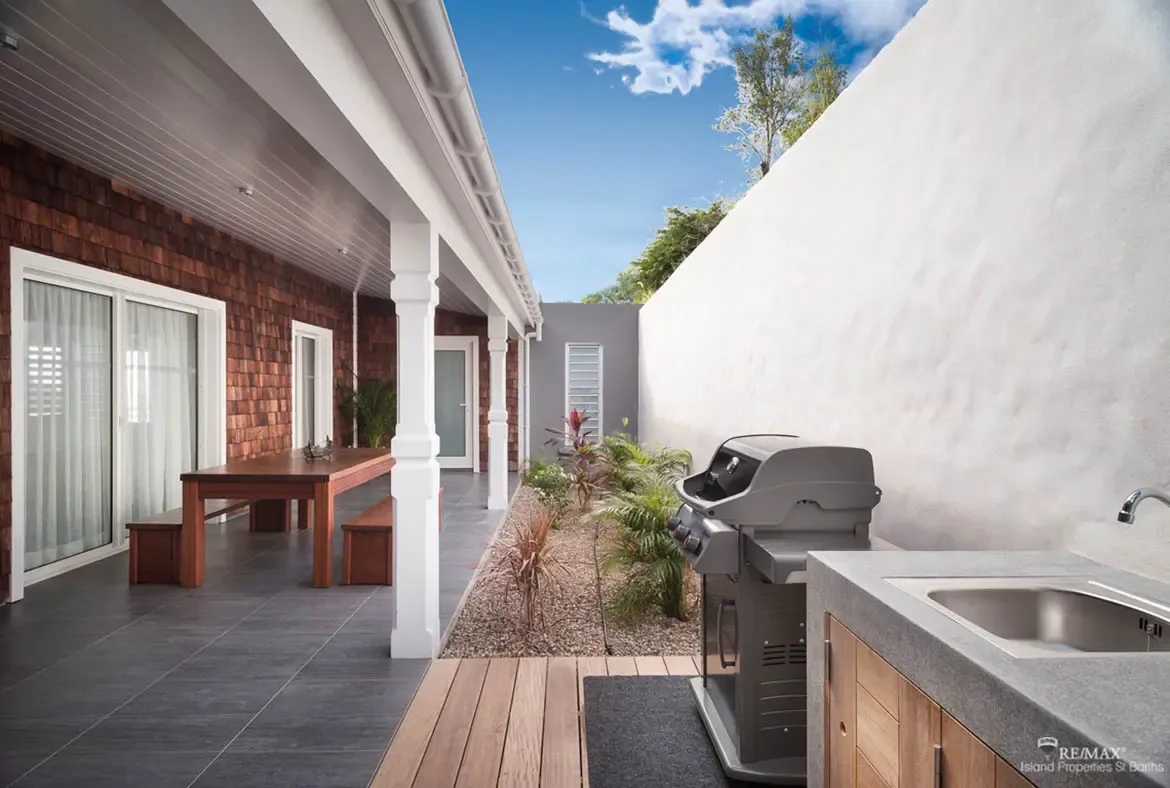 A wooden dining table with benches sits under a covered porch with white columns beside a stainless steel grill and stone countertop outdoor kitchen. Red-leafed tropical plants and palm foliage line the gravel garden strip between—a chef's outdoor paradise for al fresco dinners.