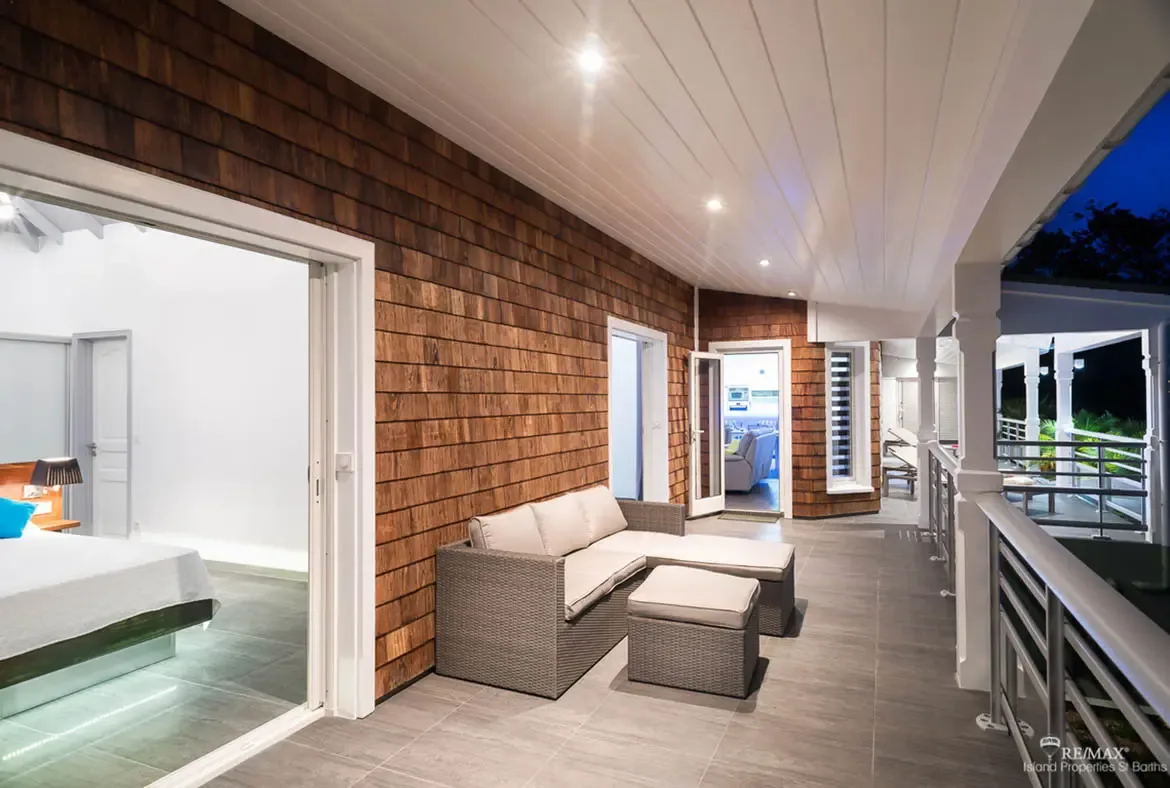 A wicker sectional with beige cushions and matching ottomans sits beneath a white paneled ceiling on sleek gray tiles. Warm wooden shingles line the wall while open doors reveal a bedroom with white linens and a blue accent pillow—indoor-outdoor living at its Caribbean finest.