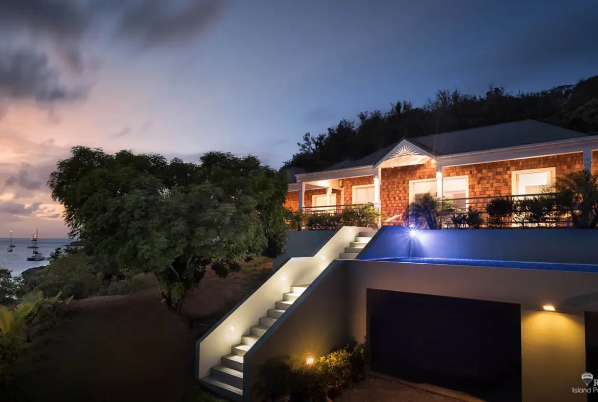 A warm-shingled villa glows with exterior lighting as dusk settles over the twilight sky in soft pink and blue. Lit stairs ascend beside a pool with an underwater blue glow while a grand tree silhouettes against sailboats on the distant ocean—evening at Villa Pleasure is pure enchantment.