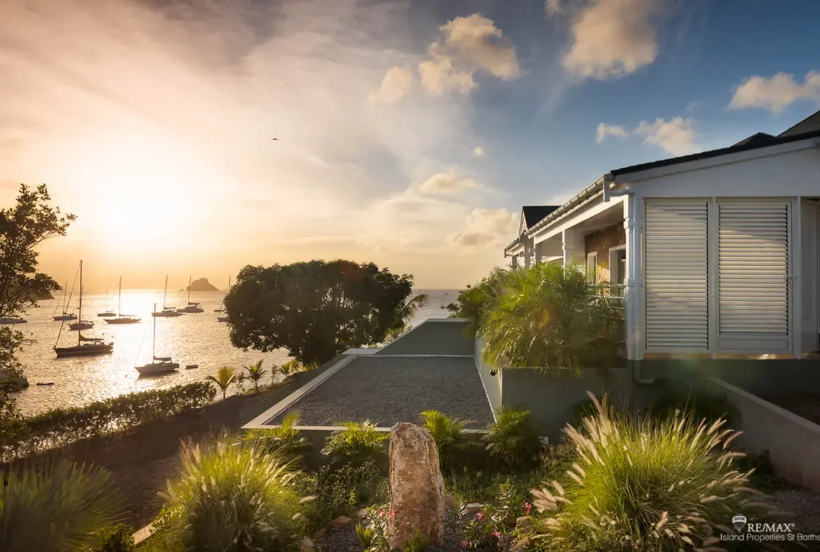A white villa with classic shutters and a covered porch perches above a bay where sailboats rest at anchor beneath a golden sunset. Flowering tropical plants frame the view as the sun dips low and paints the water in warm amber light—this is the St. Barths dream.